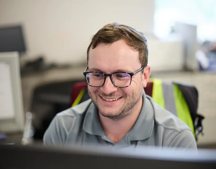 Happy employee working at a desk in an office environment, wearing glasses and a gray shirt.
