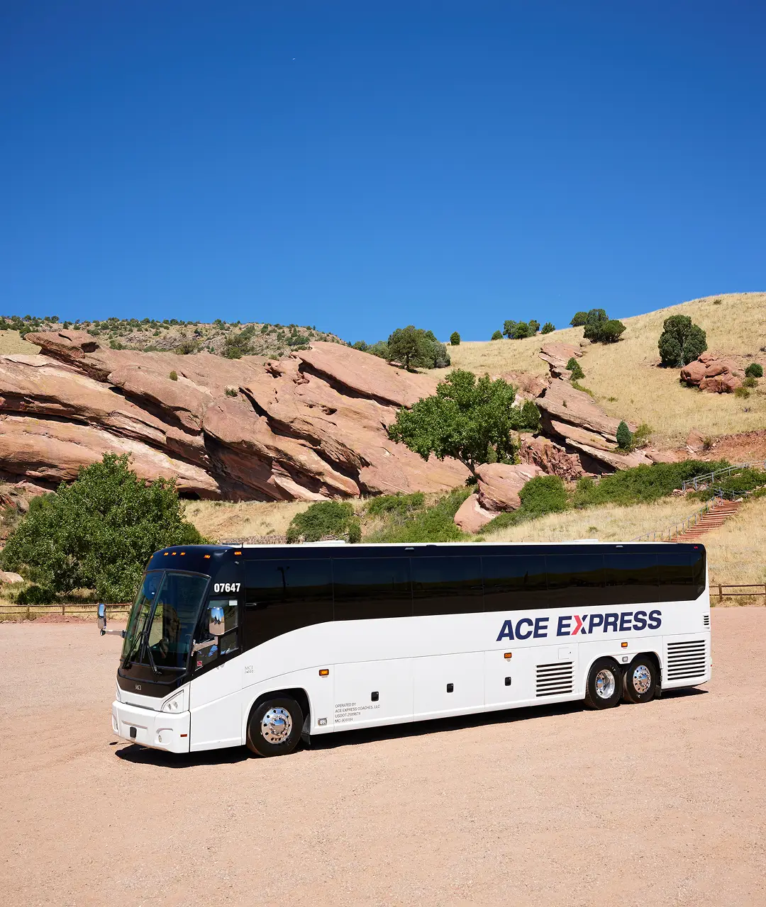Motorcoach from Ace Express Coaches parked on a dirt area with rocky hills and sparse trees under a clear blue sky.