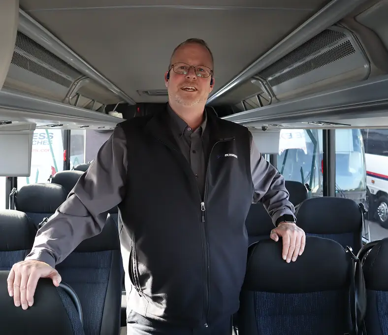 Male motorcoach driver standing inside the motorcoach, smiling, with seats and windows in the background.