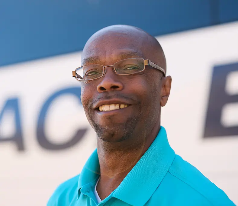Smiling male motorcoach driver wearing glasses and a blue uniform in front of Ace Express Coaches signage.