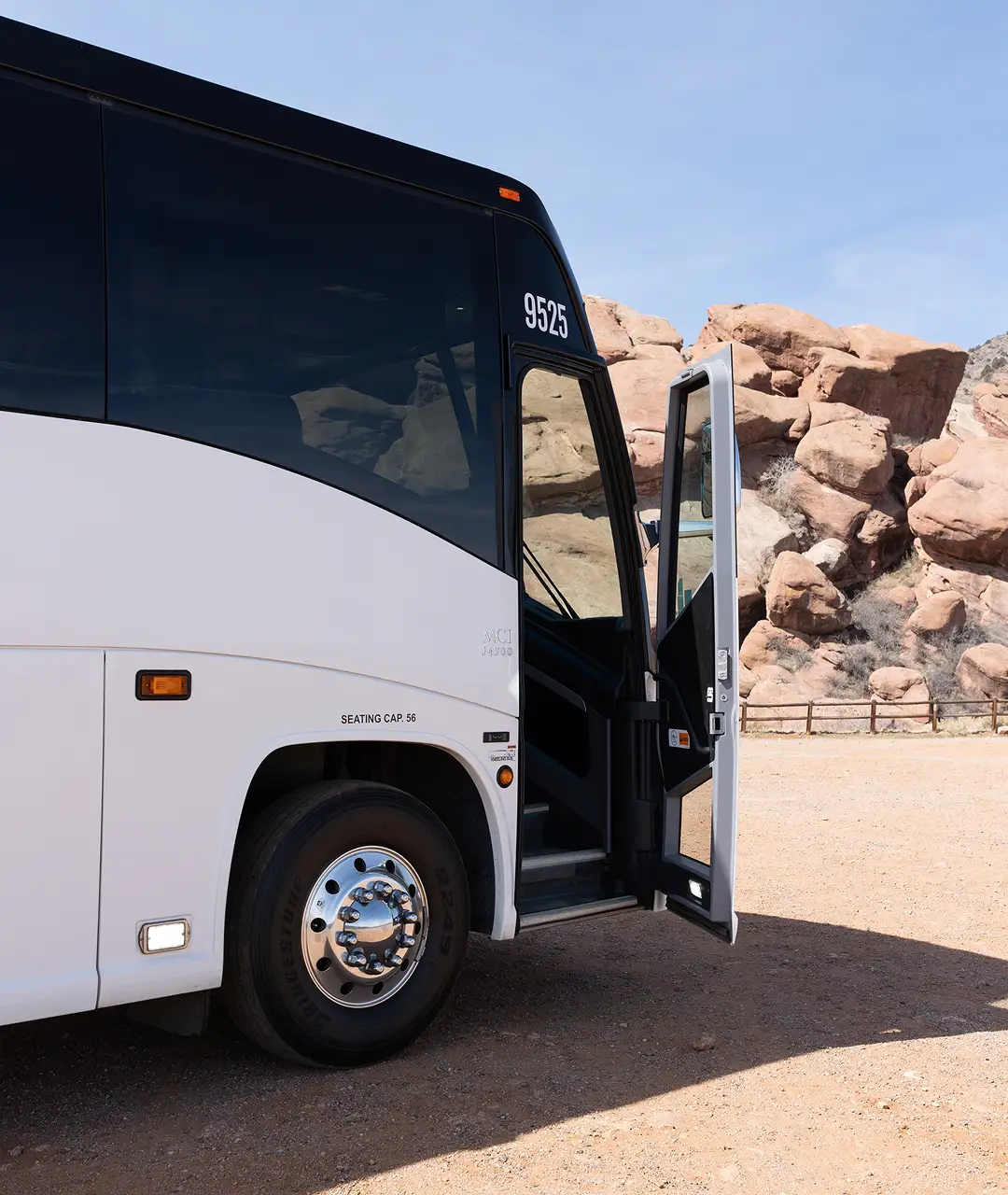 White motorcoach with open door parked in desert with rocky formations in background.
