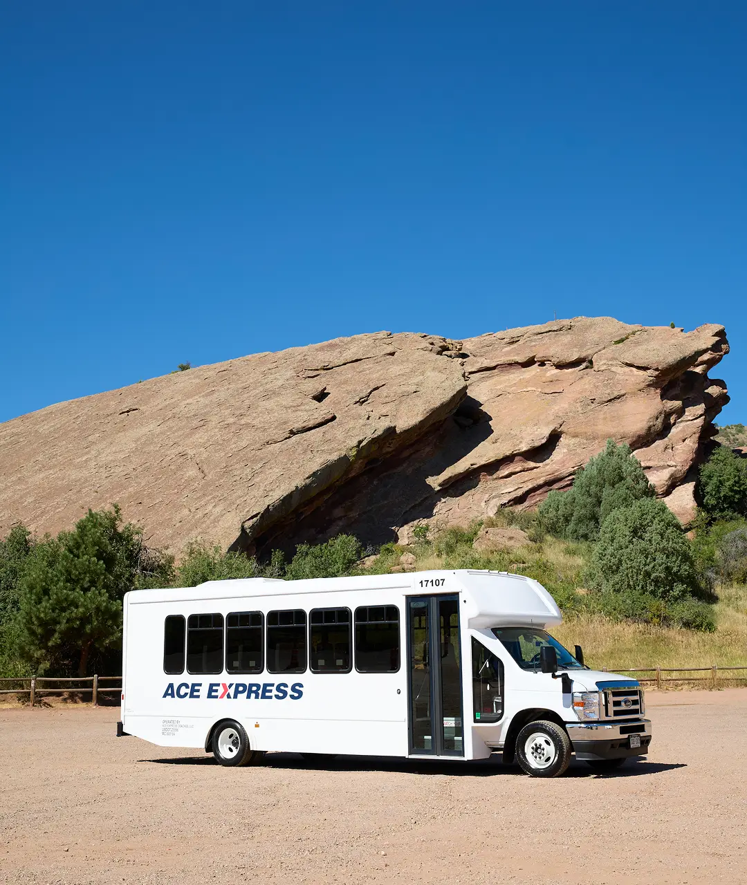 White Ace Express motorcoach parked on dirt road with rocky hills and greenery in the background.