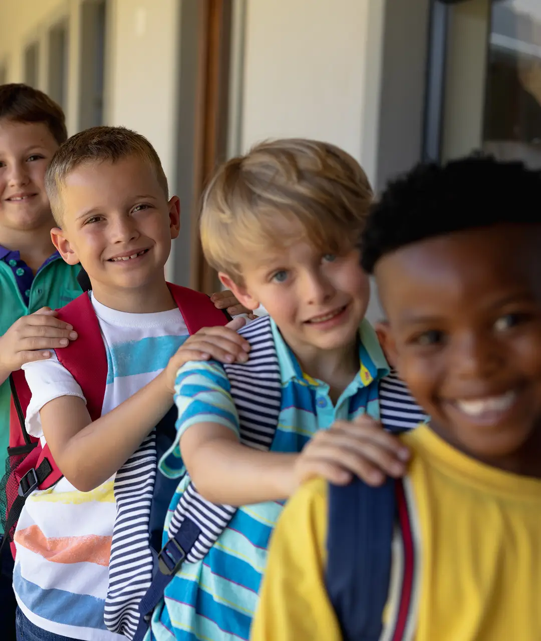 Group of happy children with backpacks with their hands on each others' shoulders in a line outside a school building