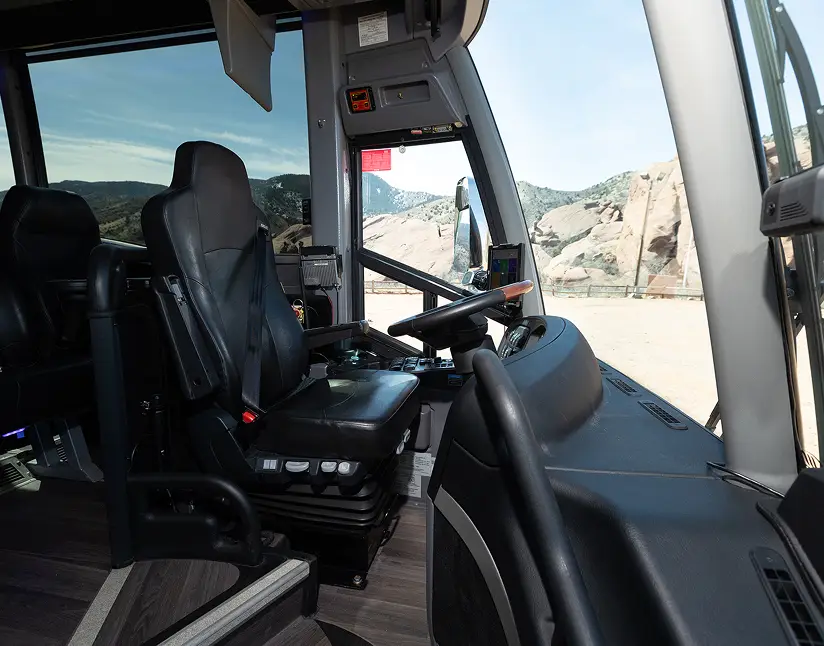 Interior view of an Ace Express Coaches motorcoach driver’s cabin with steering wheel, driver seat, dashboard controls, and panoramic front view.