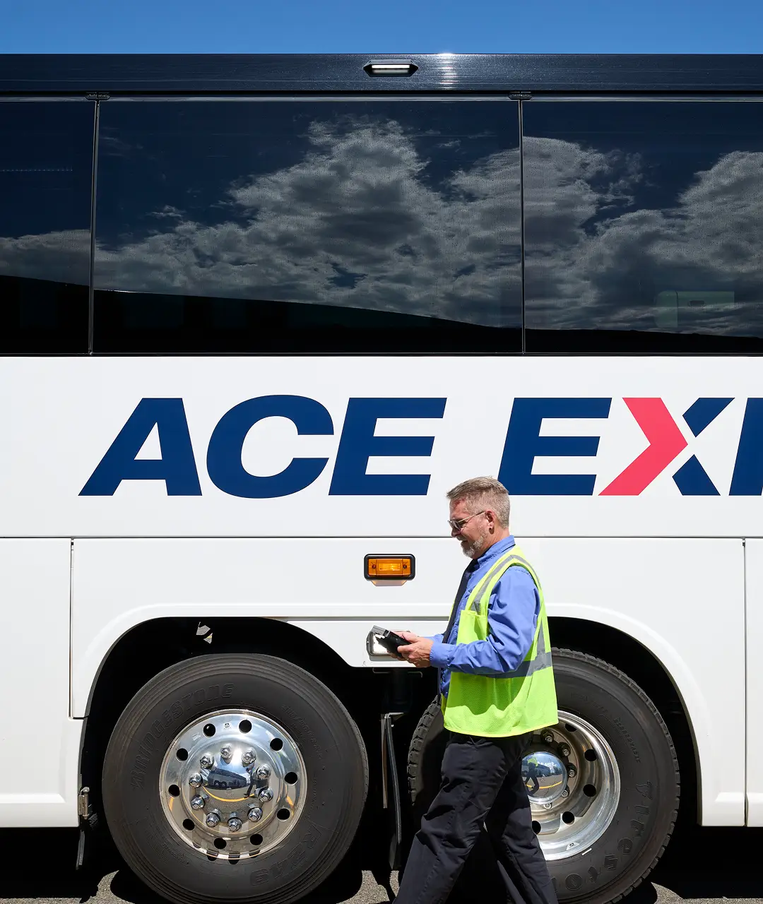 Man in safety vest using a tablet beside an Ace Express Coaches motorcoach.