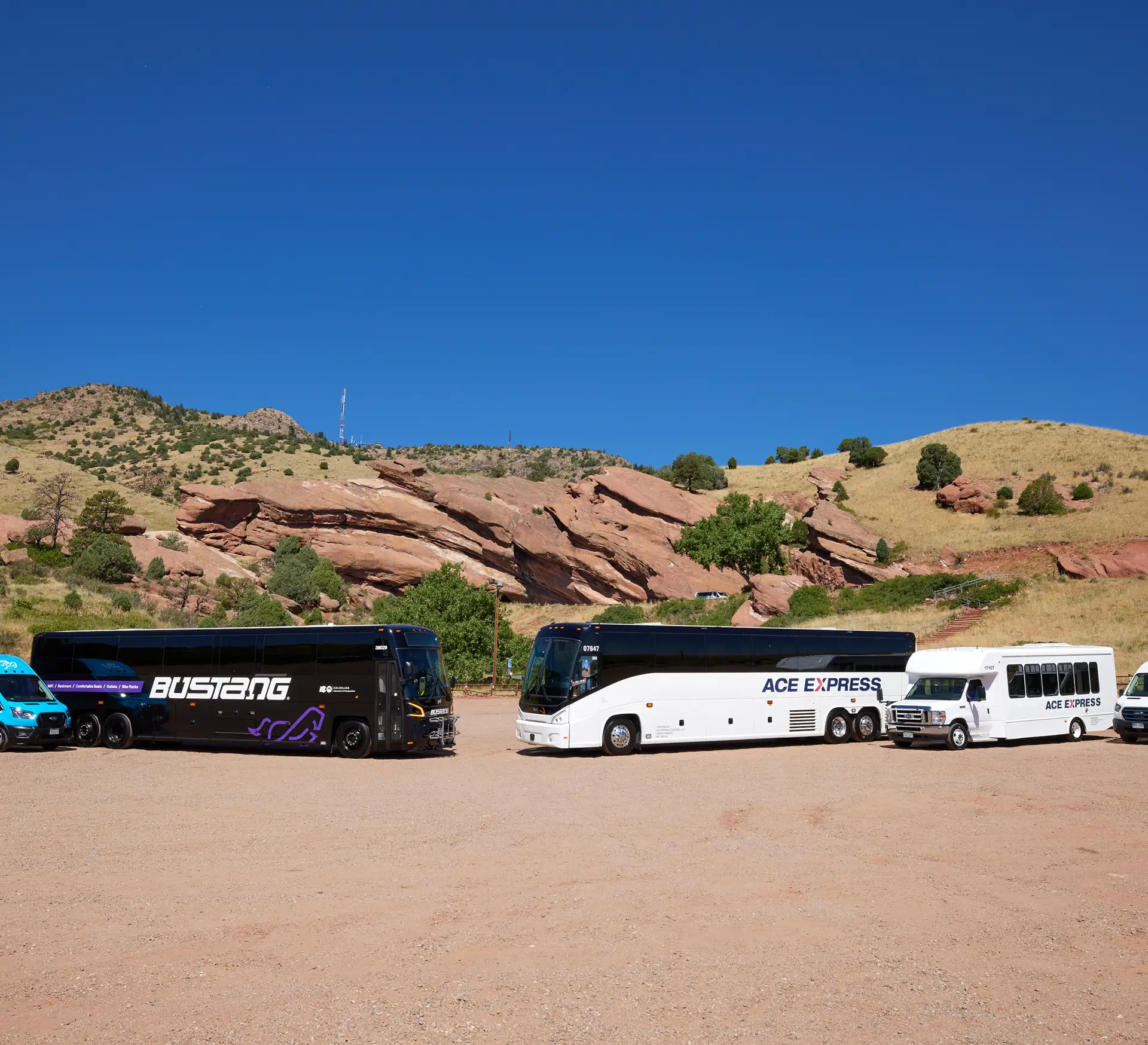 Group of motorcoaches parked on a dirt lot with rocky hills in the background.