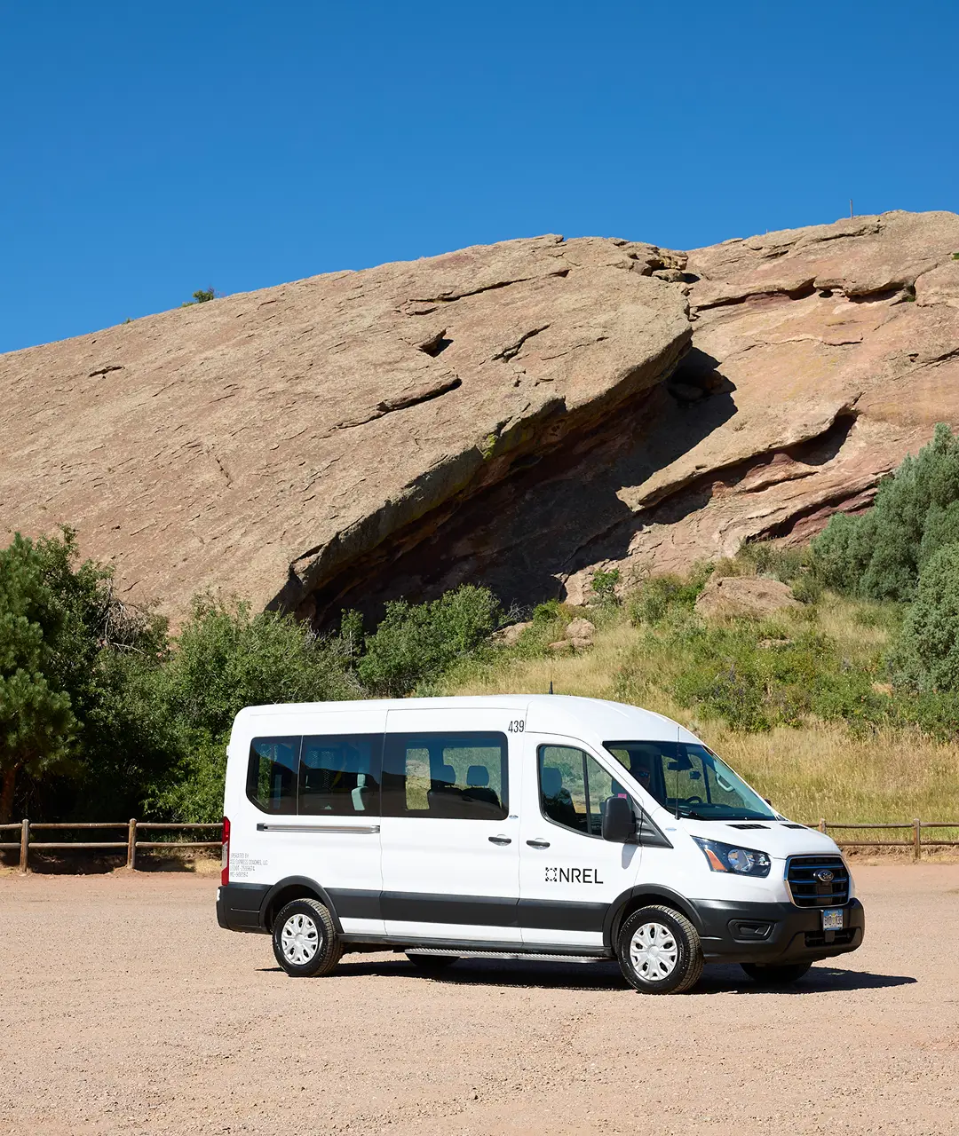 White minibus parked on dirt ground with rocky hill and greenery in background.