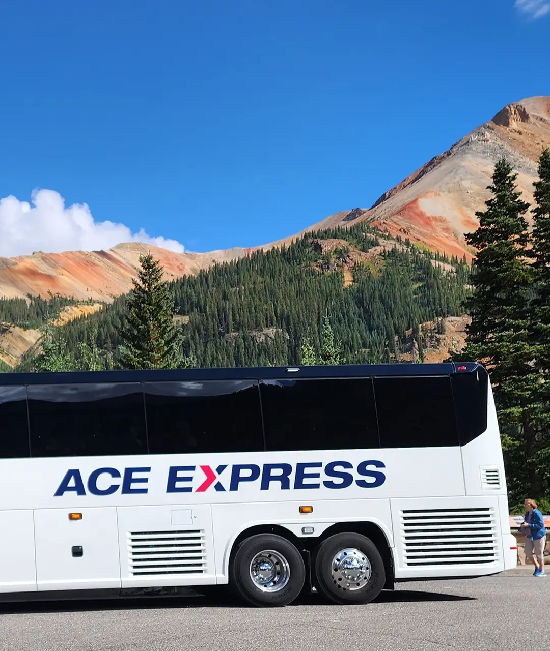 Modern white motorcoach with Ace Express Coaches logo parked near colorful mountain landscape.