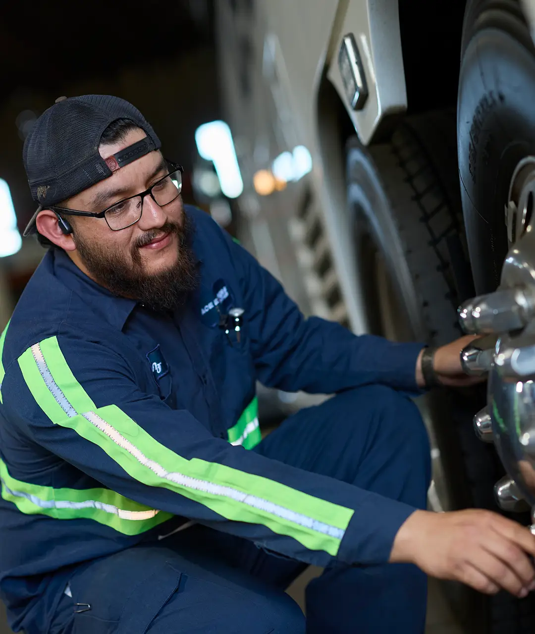 Bus mechanic inspecting a large bus tire at night, wearing safety gear and uniform.